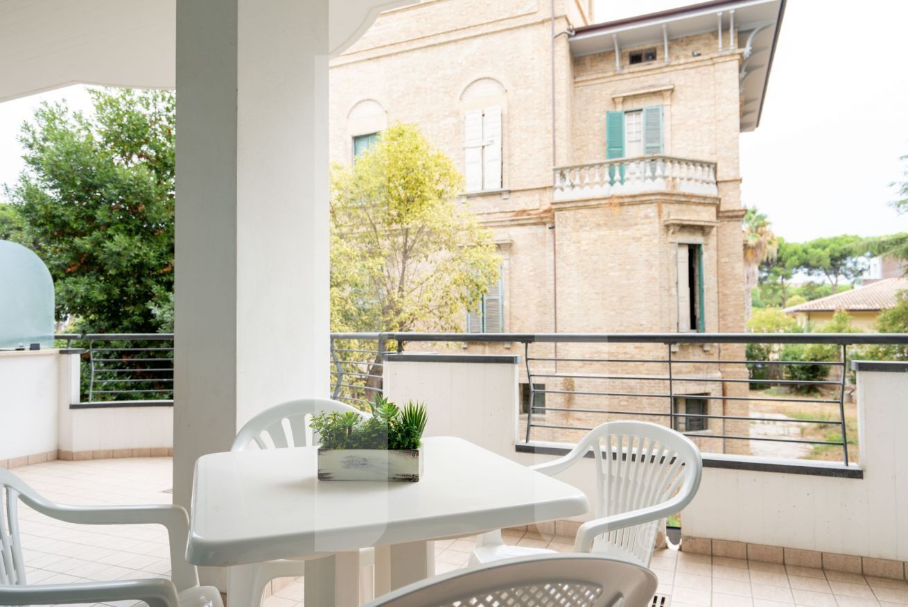 En balkon med et hvidt bord og stole, en potteplante og udsigt til en murstensbygning med vinduer og skodder.