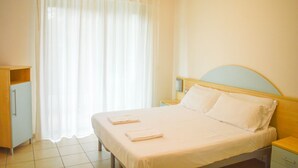 Una habitación de hotel con una cama, una mesita de noche, un armario y una ventana con cortinas translúcidas.