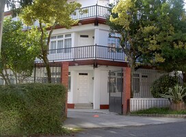 Un edificio de dos pisos con fachada blanca, un balcón con barandillas negras y un acceso con portón.