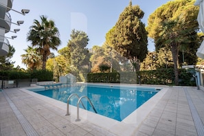 A swimming pool with a tiled deck, surrounded by trees and a clear sky.