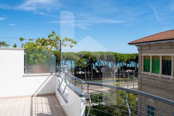 A balcony with a metal railing overlooking a green landscape and a building with a tiled roof.