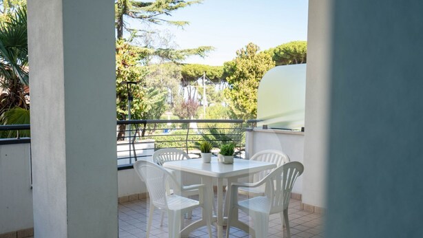 Ein Balkon mit einem weißen Tisch und Stühlen, einem Blumentopf und Blick auf Bäume und Gebäude.