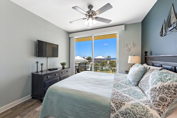Quarto com uma cama grande, um ventilador de teto, uma TV sobre um penteadeira e uma varanda com vista para edifícios.