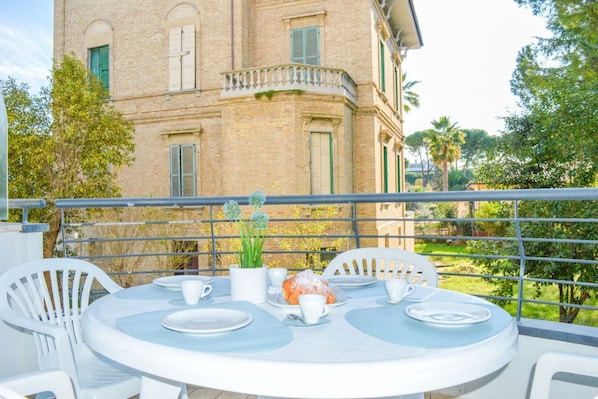 A round table with chairs and a dining setup on a balcony with a view of a building and greenery.