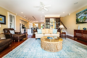A spacious living room with a large round coffee table, a sofa, and a leather armchair. There is a ceiling fan, a staircase leading to another level, and a kitchen in the background.