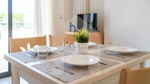 Uma mesa de jantar posta com pratos, talheres e um copo, com uma planta sobre a mesa e uma televisão ao fundo.