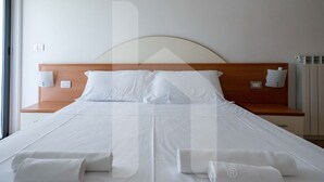 Een hotelkamer met een groot bed, witte lakens, twee nachtkastjes met lampen en uitzicht op buiten.