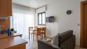 Een moderne woonkamer met een bank, een eettafel met stoelen, een aan de muur bevestigde televisie en een keuken met fornuis en kastjes.