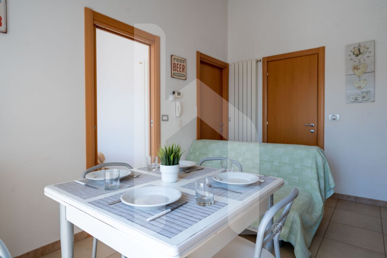 Une petite pièce, bien rangée, avec une table à manger dressée pour deux personnes, un lit recouvert d’une couverture verte et une porte sur laquelle est affiché un panneau indiquant « Bière gratuite ».