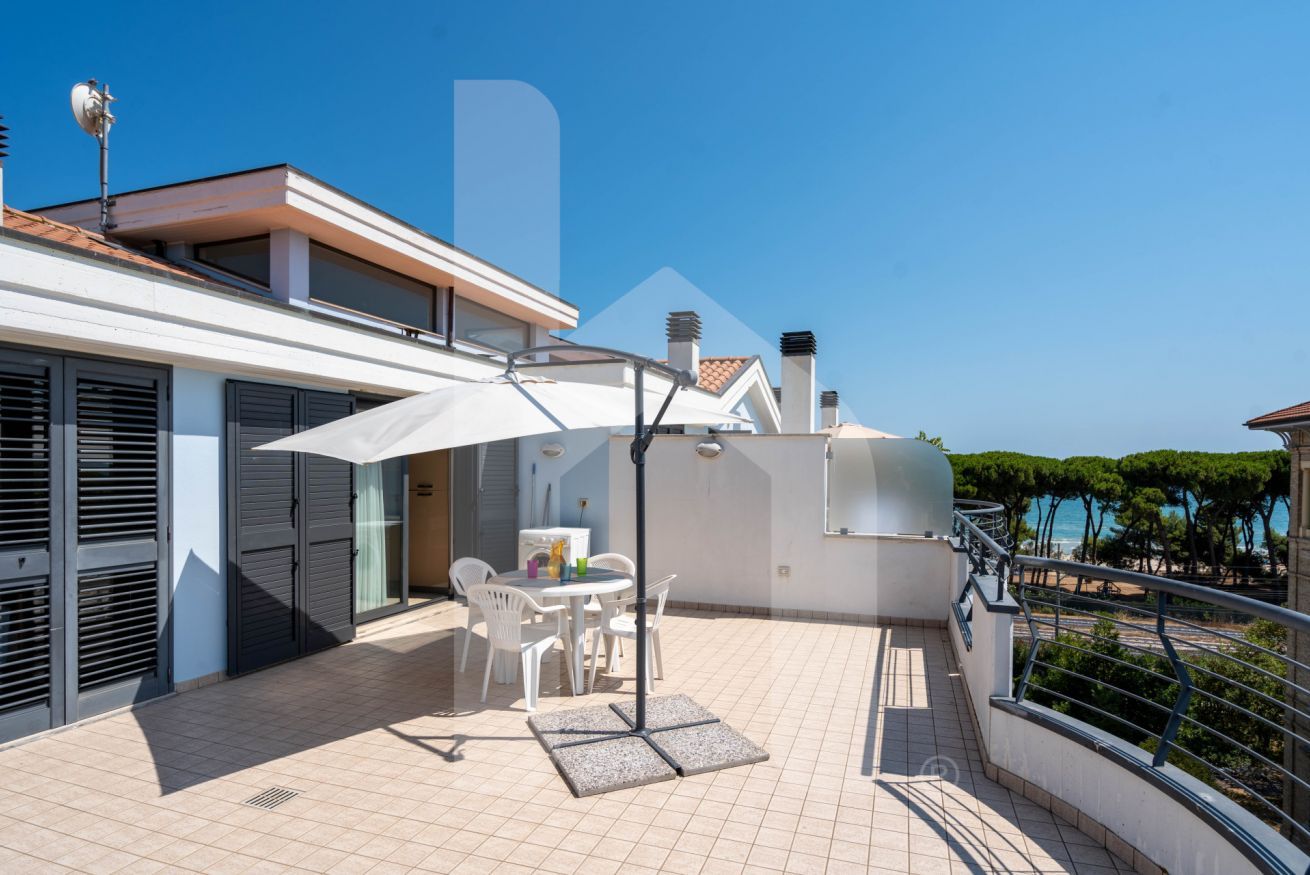 A modern outdoor patio with a table and chairs, a white umbrella, and a view of the sea.