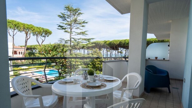 Ein Balkon mit rundem Tisch für vier Personen, umgeben von weißen Stühlen, mit Blick auf einen Swimmingpool und viel Grün.