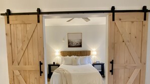 Un dormitorio con una amplia puerta de madera estilo granero, una cama con ropa de cama blanca, dos mesitas de noche con lámparas y un ventilador de techo.