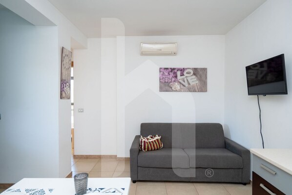 Een woonkamer met een grijze bank, een flatscreen televisie en een luchtverkoeler aan de muur.