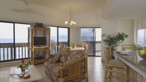 Un'ampia zona giorno con una grande finestra vista mare, una televisione su un mobile in legno, un tavolo da pranzo con sedie e un angolo cottura con un bancone e sedute.
