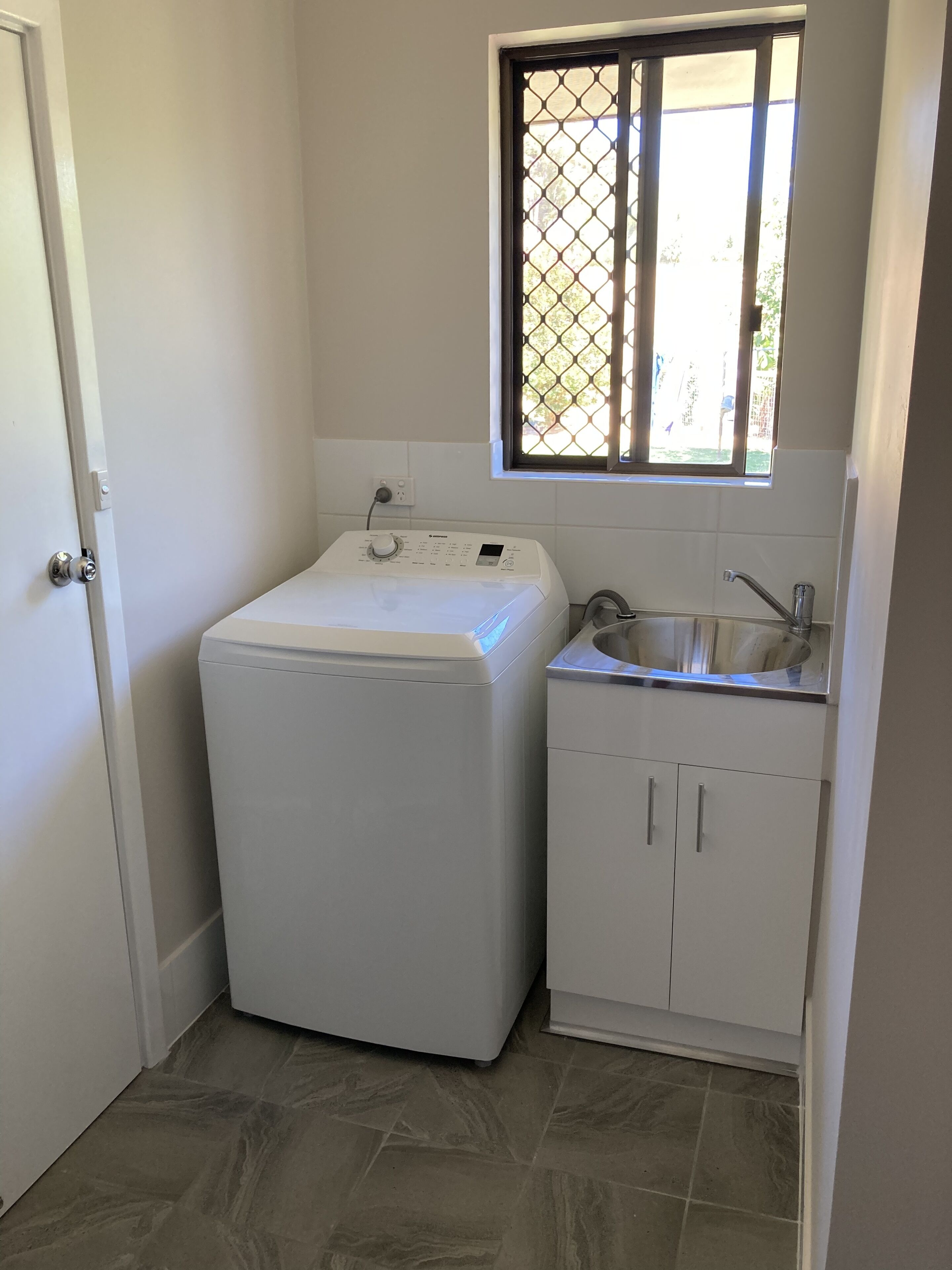 A compact laundry room with a washing machine, a sink, and a window with a metal screen.
