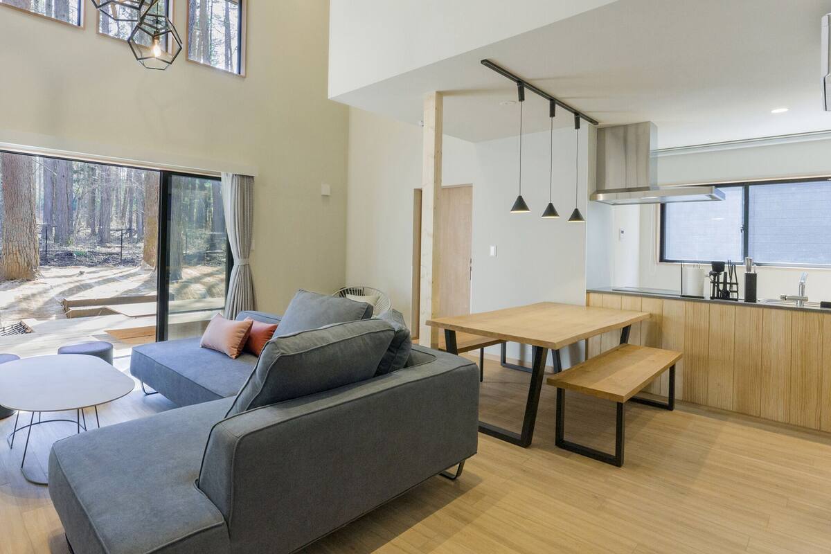 A modern living room with a grey sofa, a small round table, and a large window overlooking a forest.