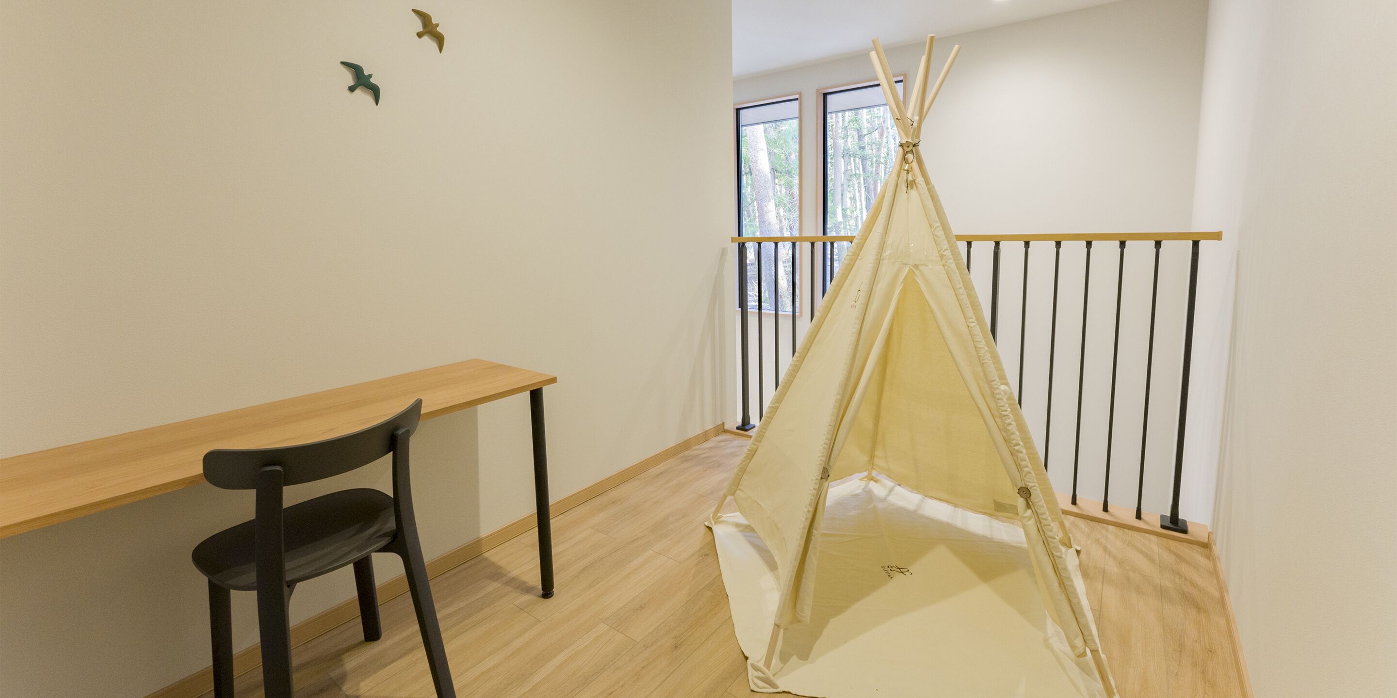 Une pièce avec un bureau en bois, une chaise noire, une structure de couleur beige ressemblant à une tente, et une fenêtre donnant sur des arbres.