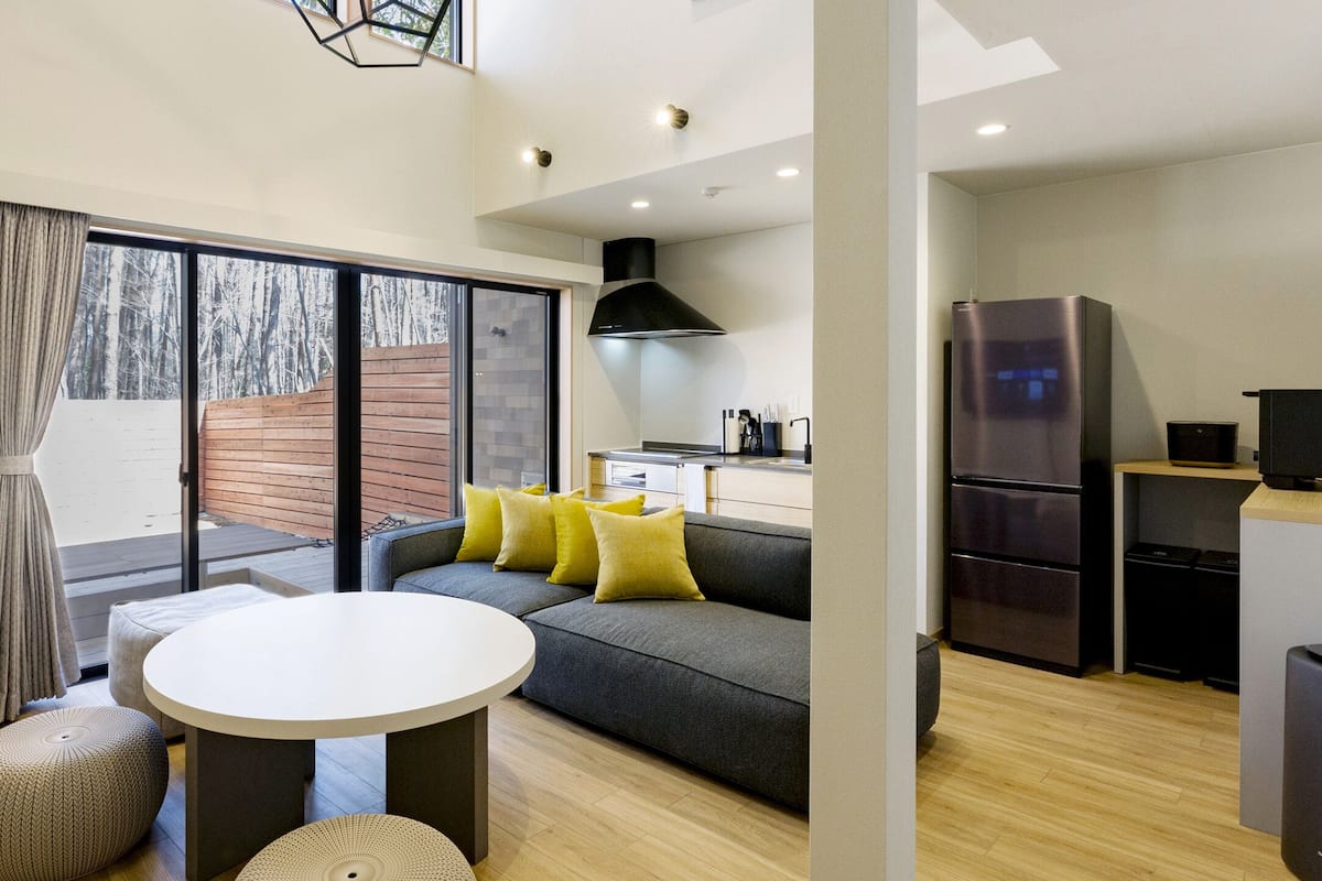 A modern living room with a grey sofa, round white table, and yellow cushions. A kitchen area with a black refrigerator and wooden flooring is visible.