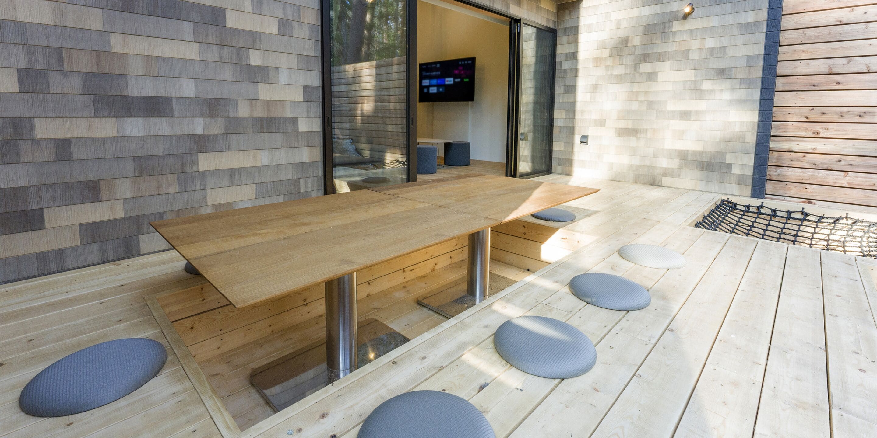 A modern outdoor dining area with a wooden table and chairs, a TV mounted on the wall, and a wooden deck.