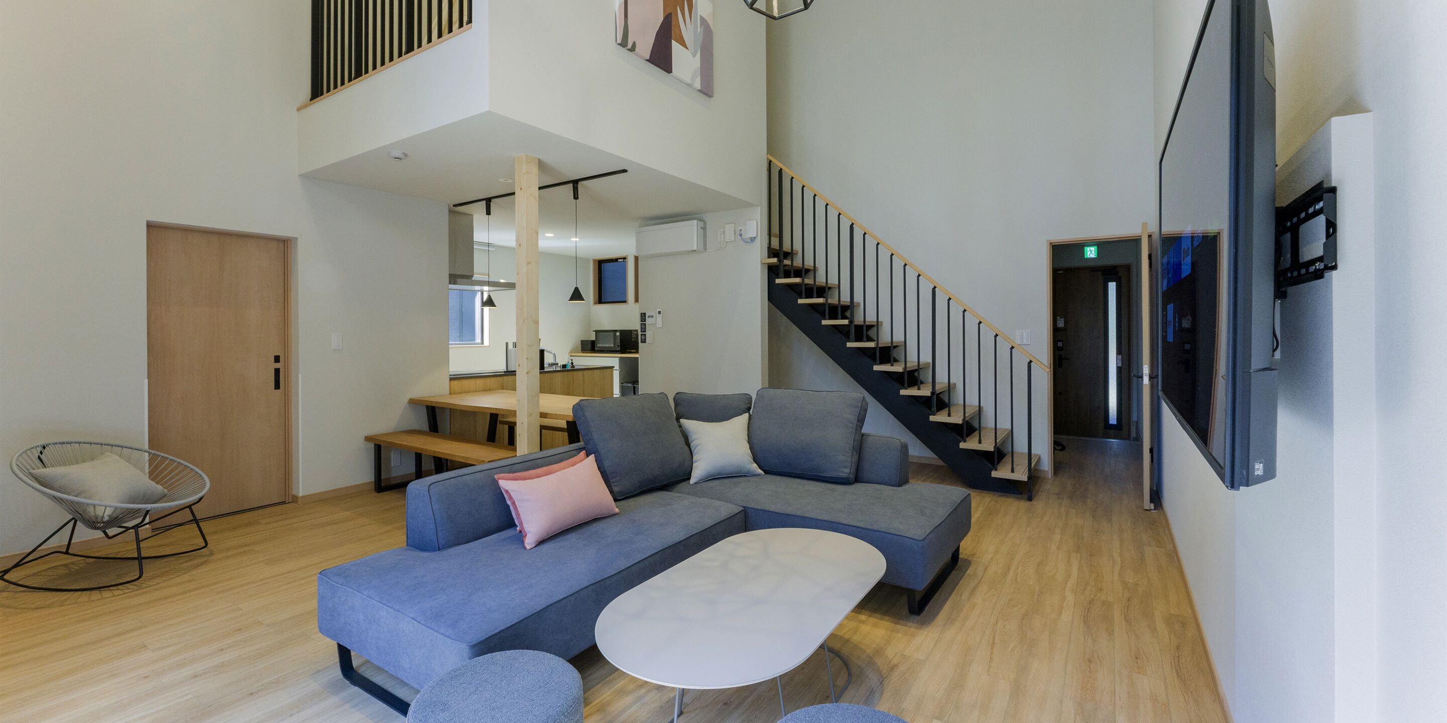 A modern living room with a sectional sofa, a round coffee table, and a dining area with a wooden table and chairs. A staircase with a wooden railing leads to an upper level.