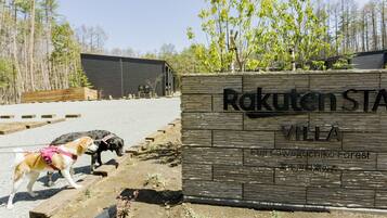 Een houten bord met de tekst 'Rakuten Stay Villa' en 'Fujikawaguchiko Forest', een hond aan de lijn, en een bebost landschap op de achtergrond.