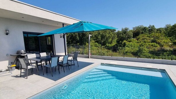 Un área de piscina exterior moderna con una piscina azul cristalina, una zona de barbacoa y una mesa de comedor con sillas.
