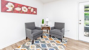 Ein Zimmer mit zwei grauen Sesseln, einem gemusterten Teppich, einem floralen Gemälde in Rot, einem kleinen Tisch mit Lampe und einer zur Außenseite führenden Tür.