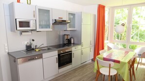 A modern kitchen with a built-in microwave, oven, and coffee maker. There is a dining area with a table and chairs, and a window with curtains.