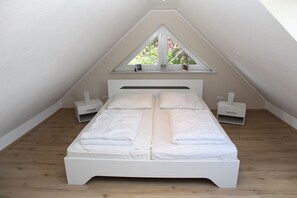 Ein Schlafzimmer mit schrägverdachter Decke, einem Bett mit weißer Bettwäsche, zwei Nachttischen und einem Fenster mit Blick auf Grünflächen.