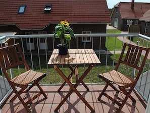 Eine Holzterrasse mit einem Tisch, zwei Stühlen, einem Topf mit einem Sonnenblume und Blick auf ein Backsteinhaus mit rotem Dach.