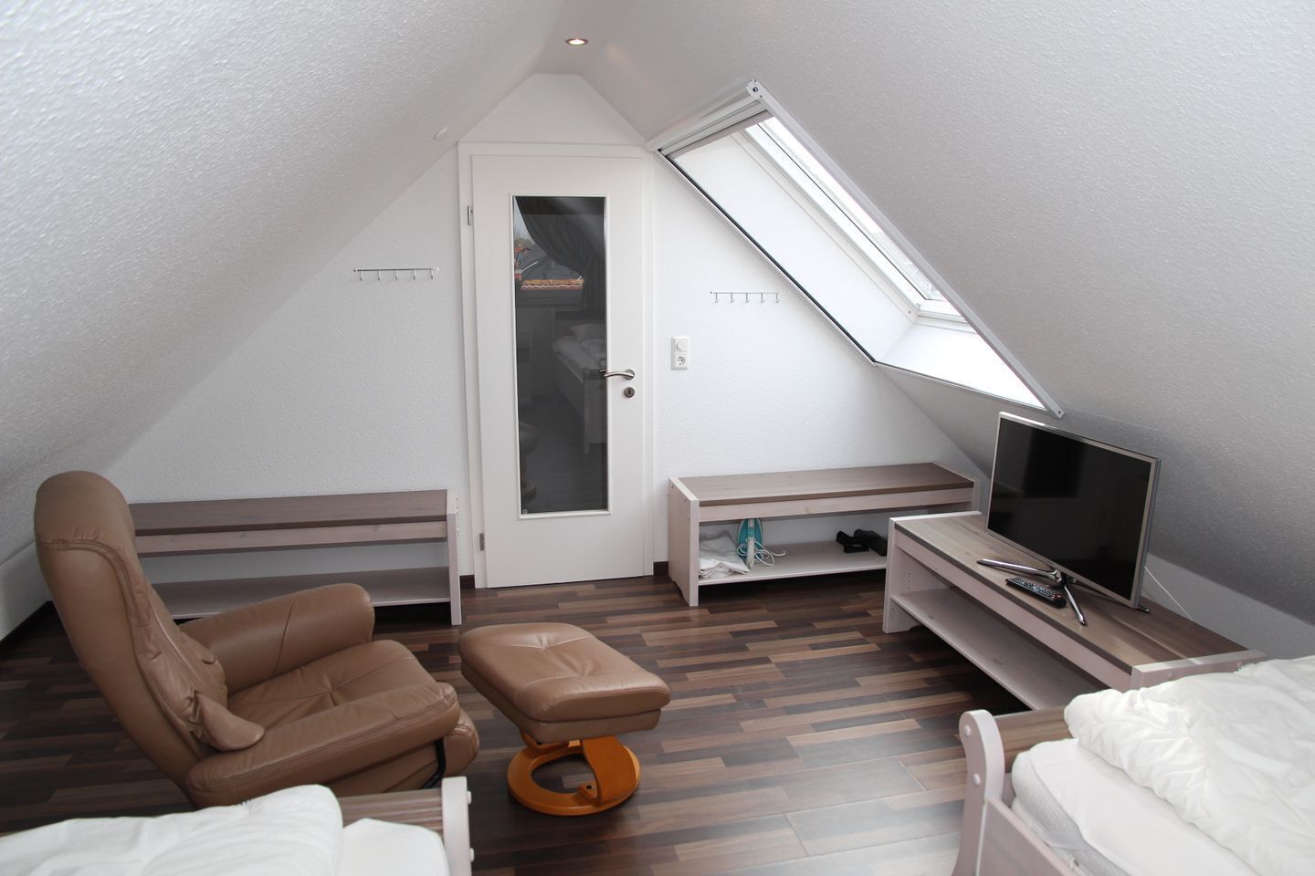 Ein kleiner, gut beleuchteter Raum mit schrägem Dach, einem Einzelbett, einem braunen Ledersessel, einem kleinen Tisch mit Fernseher und Fernbedienung sowie einer Tür mit Milchglasfenster.