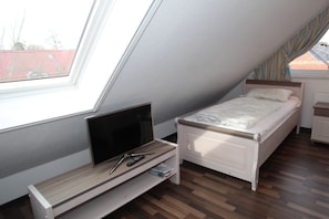 Een kleine, goed verlichte kamer met schuine wanden, een enkel bed, een televisie op een houten standaard en een raam met uitzicht op gebouwen.