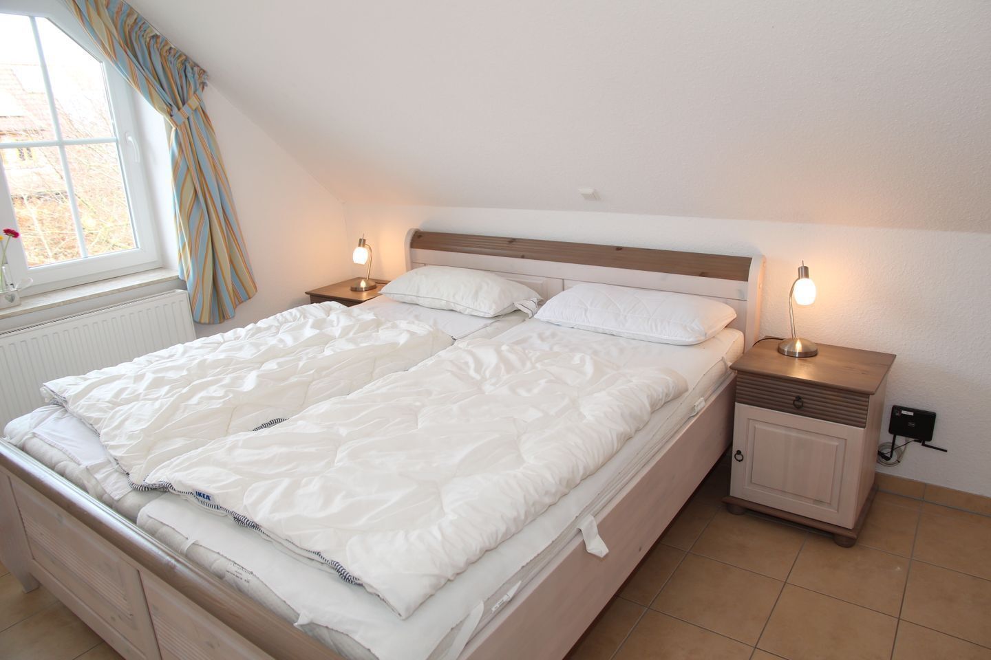 Una cama doble con ropa de cama blanca, dos mesitas de noche con lámparas, una ventana con cortinas y un radiador.
