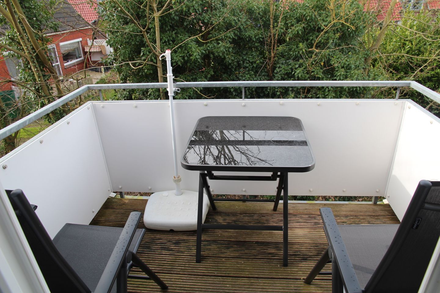 A balcony with a small table and two chairs, a white railing, and a view of greenery and a building in the background.