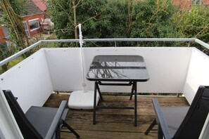 Ein Balkon mit einem kleinen Tisch und zwei Stühlen, einem weißen Geländer sowie Blick auf Grünflächen und ein Gebäude im Hintergrund.