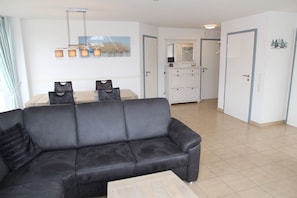Ein modernes Wohnzimmer mit einem schwarzen Sofa, einem Esstisch mit Stühlen, einer Küchenzeile mit Schränken und einem großen Fenster mit Vorhängen.