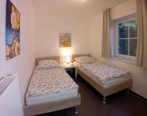 Una habitación pequeña con dos camas, una mesita de noche con lámpara, un cuadro en la pared y una ventana con cortinas.