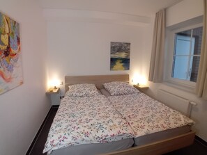 A single bed with a floral bedspread, two bedside lamps, a painting on the wall, and a window with curtains. - Hummel Hummel - Dog-friendly vacation apartment (70sqm) for 4 people with garden (Krummhörn)