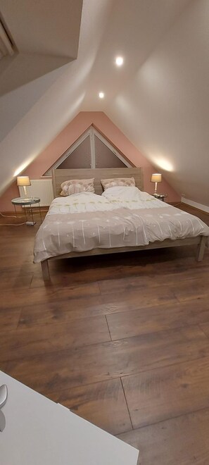 Una camera da letto con soffitto spiovente, un letto con copripiumino bianco, due comodini con lampade e un pavimento in legno.