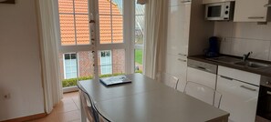 Una cucina con mobili bianchi, un forno a microonde e un lavello. Si vede una finestra con tende e un tavolo con sedie.
