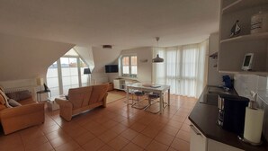Un apartamento moderno con sala de estar, comedor y cocina. Hay un sofá, una mesa de comedor con sillas, una encimera de cocina y un televisor montado en la pared.
