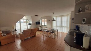 Eine moderne Wohnung mit Wohnzimmer, Essbereich und Küche. Es gibt ein Sofa, einen Esstisch mit Stühlen, eine Küchenarbeitsplatte und ein an der Wand befestigtes Fernsehgerät.