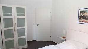 Une chambre avec un lit, une armoire blanche aux portes en verre dépoli, une porte blanche, une petite table de chevet avec une lampe et un tableau encadré au mur.