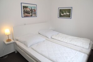 A single-bed room with white bedding, a bedside table with a lamp, and two framed pictures on the wall.