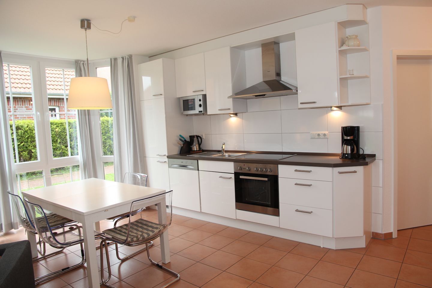 A modern kitchen with white cabinets, a stainless steel oven, a microwave, and a coffee maker. There is a dining table with chairs and a window with curtains.