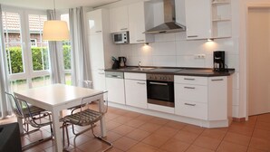 Une cuisine moderne avec des meubles blancs, un four en acier inoxydable, un four à micro-ondes et une machine à café. On y trouve une table à manger avec des chaises et une fenêtre avec des rideaux.