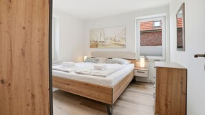 A bedroom with a bed, bedside tables, a nightstand, a wooden cabinet, a painting, and a window with a view of a brick building. - Strandfloh - Gemütliche Ferienwohnung mit Fahrstuhl (Norden)