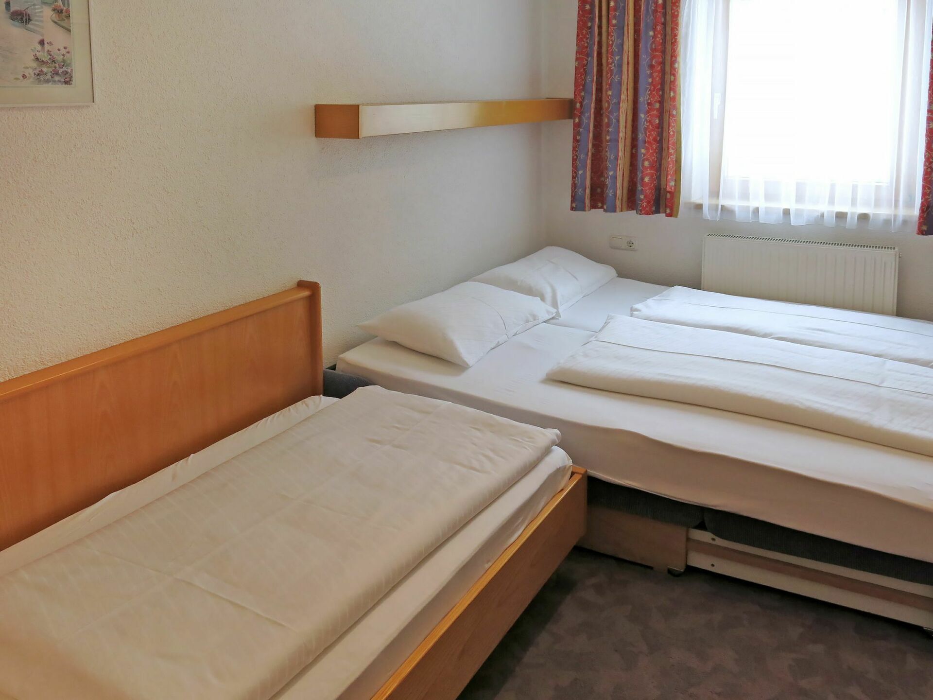 Ein Zimmer mit zwei Einzelbetten, einem Holzkopfteil, einer kleinen Ablagefläche und einem Fenster mit Vorhängen.