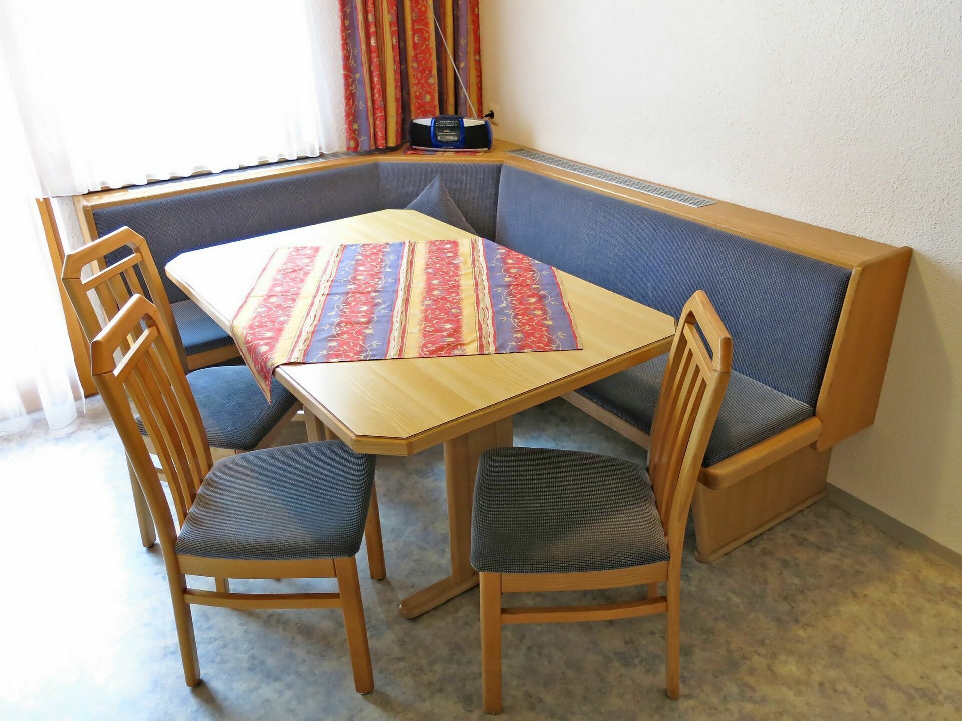 A corner dining area with a wooden table, two chairs, and a bench with a patterned tablecloth.