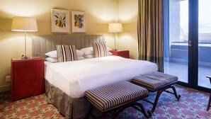 A hotel room with a large bed, two bedside tables, a bench, and a view of the sea through a glass door.